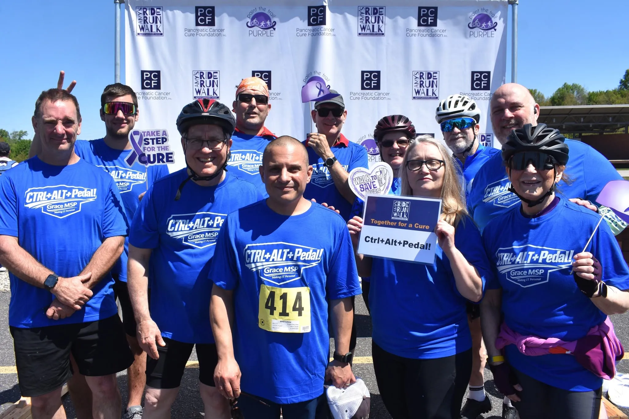 The GraceMSP Amy’s Ride team in matching blue shirts in front of the event backdrop, holding a Ctrl+Alt+Pedal! sign.